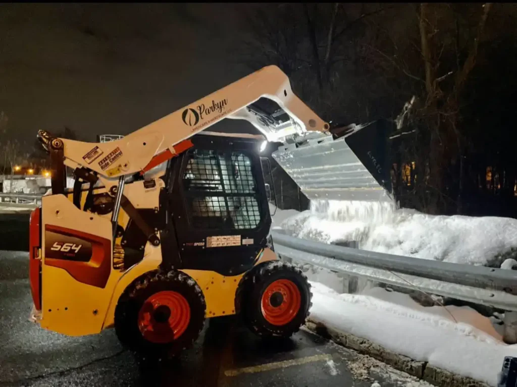 A compact skid-steer loader with a raised snow bucket is clearing a large pile of snow beside a roadside guardrail at night.