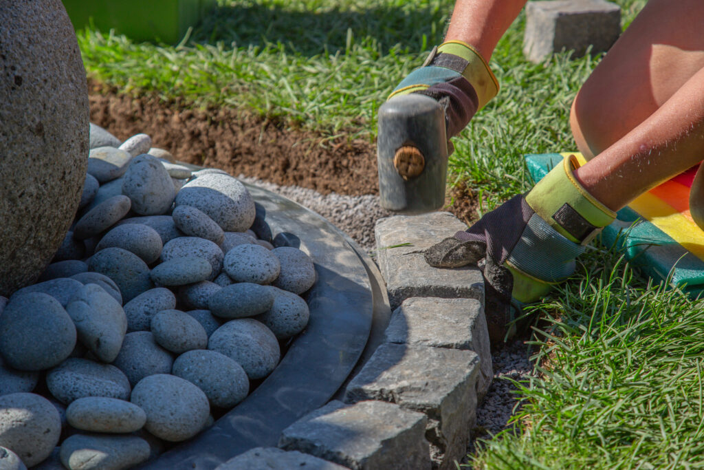 Worker installing stone landscape edging around decorative river rocks and grass border in a residential yard